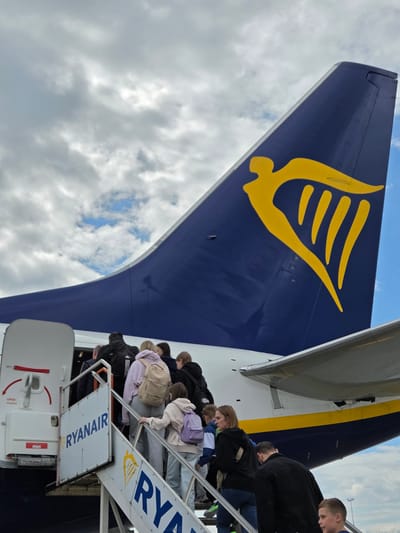 In Balice, Poland, a group of passengers, including adults and children, ascend a portable staircase to board a Ryanair airplane. The tail of the aircraft, featuring the distinct yellow harp logo on a navy blue background, is prominently visible against a cloudy sky. The side of the plane displays the airline's name, "RYANAIR," in large blue letters on a white panel affixed to the staircase. Passengers are seen from behind and in profile, carrying backpacks and dressed in casual attire, suggesting they are preparing for travel. The scene captures the common activity of boarding an aircraft at an airport.
