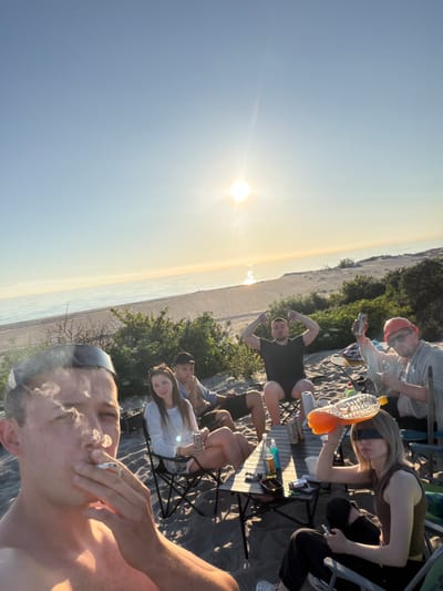 The image depicts a group of young adults gathered on a sandy beach near the city of Povarovka, Russia. The sun is setting over the calm sea, casting a warm glow on the scene. In the foreground, a young man is captured in a close-up, smoking a cigarette with smoke billowing around his face. He is wearing a black visor and appears to be topless.
Behind him, seated in folding chairs on the sand, are several other individuals. A young woman with long brown hair is looking towards the camera, holding a can. Next to her, a man in a dark t-shirt and shorts is seated, with his arms raised in a gesture. Further back, another man is visible, wearing a red cap and sunglasses, holding up a can and a bottle. A table is set up between them, holding various drinks and other items. The background shows the beach and vegetation, with the expanse of the sea and sky. The overall atmosphere appears casual and relaxed, suggesting a social gathering during sunset.