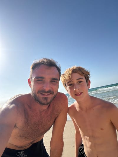The image displays two males, an adult and a juvenile, posing for a photograph on a beach in Beit Yanai, Israel. The adult male, on the left, has dark hair, a beard, and is shirtless, wearing black swim trunks. The juvenile male, on the right, has reddish-brown hair and is shirtless, with dark swim trunks partially visible. Both individuals are smiling and looking directly at the camera. The background consists of light-colored sand, the blue-green ocean with small waves, and a clear blue sky. A bright sun glare is present in the upper left portion of the sky.