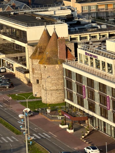 This is an aerial, eye-level, long shot of a city intersection in Dieppe, France, with a focus on the Mercure Hotel and a historic stone tower. The Mercure Hotel is a modern building with a glass facade on the upper floors displaying the hotel's name in purple lettering. The lower floors are clad in grey tiles with numerous windows. Purple banners reading "Mercure HOTEL" hang on the exterior. In front of the hotel is an entrance with a brown awning, planters with vegetation, and outdoor seating visible through the glass. A white bicycle is parked near the entrance.
To the left of the Mercure Hotel is a historic, round stone tower with two conical, moss-covered roofs, resembling a castle turret. Adjacent to the tower and to the left of the hotel is a building with the word "CASINO" in large letters on its facade. This building has a glass exterior and a beige upper section.
The street in the foreground is paved with red brick and asphalt. A pedestrian crossing with white stripes is visible. Several streetlights with multiple lamps are positioned along the sidewalk. A car is parked on the street to the left of the intersection. Further left, several parked cars are visible in a lot or on the street. A traffic sign indicating a crossroad is present on the sidewalk. Green grass borders the street and sidewalk. The background shows rooftops of other buildings, including tiled roofs and modern structures with ventilation systems. The lighting suggests it is late afternoon or early evening, with long shadows cast across the scene.