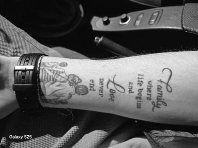 This black and white, close-up image depicts a human forearm, displaying several tattoos and a watch, positioned within what appears to be a car interior. The forearm is adorned with a black digital watch with a buckle on the wrist, beneath which a thin, braided bracelet is visible. Two primary tattoos are present: one depicting two stylized figures, possibly children, engaging in an activity, and another featuring text in a calligraphic font that reads "Family where life begins and Love never end." Visible arm hair covers the forearm. The background shows textured fabric, likely upholstery, to the left and below the arm, and elements of a car's center console, including buttons and a handbrake lever, to the upper right. A "Galaxy S25" watermark is located in the bottom left corner. The scene is located in Novo Delchevo, Bulgaria.