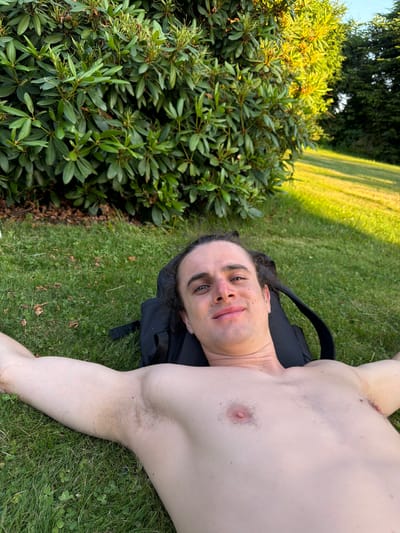 A young, shirtless person with long, dark, curly hair is lying on their back in a grassy area, with their arms outstretched. They are wearing a backpack. In the background, there is a large green bush and a lawn bathed in sunlight. The location is specified as the city of Mordarka, Poland. The scene appears to be outdoors, possibly in a park or backyard, on a sunny day.