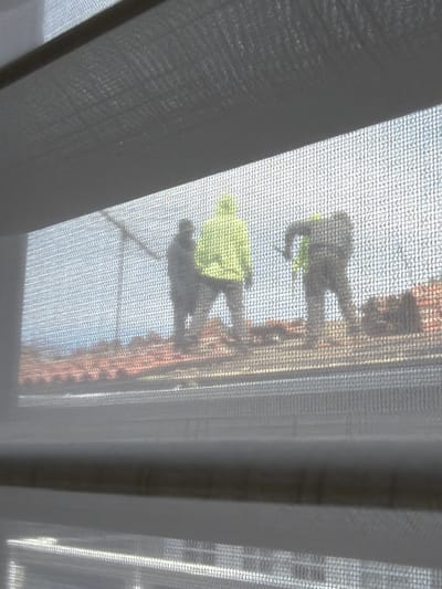 The image displays a daytime scene viewed through a translucent, light-colored mesh screen, likely a window screen or blind. Three individuals, presumably construction workers, are observed on a terracotta-tiled rooftop. The central figure wears a bright, high-visibility yellow or lime green hooded jacket and stands upright, facing slightly left. To their immediate left, a person in dark attire is partially visible, holding a long, slender object that extends upwards and to the left. To the right of the central figure, a third person, also in dark clothing, is bent over with their back partially visible, engaged in an action involving a tool held downwards towards the roof. A flash of yellow/green material is visible under their right arm. The roof features numerous rows of reddish-brown tiles. The background shows a light blue sky, suggesting clear weather. The scene depicts roofing work occurring on a building, consistent with a residential or commercial structure, located within Palos Verdes Estates, United States.