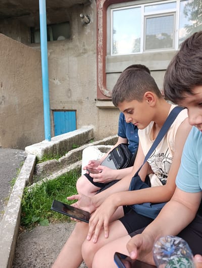 The image shows three young individuals, likely boys, sitting on concrete steps outdoors. The setting appears to be an urban environment, possibly in Rezina, Moldova, given the architectural style and the presence of a blue-painted wooden door in the background.

The person in the foreground, on the right, is partially visible and is holding a clear plastic water bottle with a blue cap. They are wearing a light blue t-shirt and dark shorts.

The central figure is a boy with short, dark hair, wearing a light beige sleeveless t-shirt with a graphic print and black shorts. He is holding a black tablet computer in his hands and looking down at it. A dark blue shoulder bag with a strap is slung across his body.

To the left of the central figure is another boy wearing a dark blue polo shirt and dark shorts. He is also holding a black tablet and looking down at it. A black bag with a silver zipper is visible on his lap.

The background consists of a textured concrete wall with a small, bright blue wooden door. A tall, thin blue pipe runs vertically along the wall. A window with a white frame is visible above and to the right of the door. Green grass and weeds grow in patches around the concrete steps. The overall impression is of a casual outdoor gathering of young people engaged with their devices.