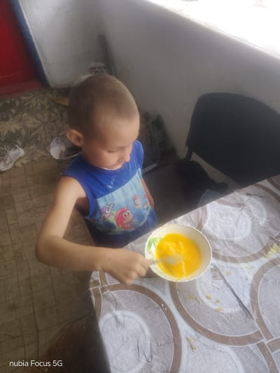 In Sestrynivka, Ukraine, a young boy with short brown hair and wearing a blue sleeveless shirt with cartoon characters is leaning over a table. He is in the process of whisking a bright yellow liquid, likely eggs, in a white bowl with a green rim using a silver whisk. Flour or a similar powdery substance is visible on his arms and shirt, and some has also scattered on the table. The table surface has a patterned design of concentric circles in shades of brown and cream, and there are also some scattered crumbs. To the left of the boy, on the floor, is a white sneaker. In the background, a dark chair is partially visible, and a white wall with a rough texture forms the backdrop. The overall scene suggests a domestic setting, possibly a kitchen or a covered outdoor area, where the child is engaged in a food preparation activity.
