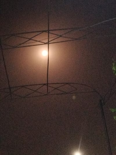 The image shows a night scene with a bright, out-of-focus light source in the upper center of the frame, possibly the moon obscured by fog or clouds, or a very bright streetlamp. Overlaid on this is a dark, angular structure made of thin beams, resembling scaffolding or part of a large tent or canopy. The background is dark and grainy, with faint hints of other lights at the bottom of the frame, suggesting an urban environment at night. The scene is likely located in the City of Türkmenabat, Turkmenistan, given the context provided. There are no people or discernible actions depicted. The overall impression is one of a hazy, dimly lit night.