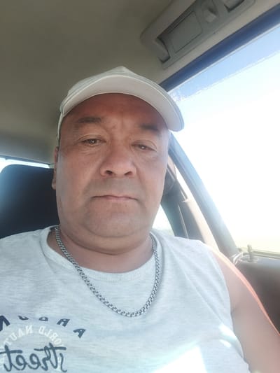 A middle-aged man with dark hair and brown eyes is shown from the chest up, looking directly at the camera. He is wearing a light-colored baseball cap, a light gray t-shirt, and a silver chain necklace. The man appears to be inside a vehicle, suggested by the car interior visible in the background, including a black car seat on the left and the car's ceiling and window on the right. The bright sunlight coming through the window indicates it is daytime. The image was taken in Bulaev, Kazakhstan.