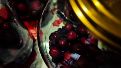 The image is a close-up, macro shot of pomegranates in a clear glass bowl, with a golden, curved object in the background. The pomegranates are a deep red color and appear to be whole, some with their skins slightly broken, revealing the arils inside. The glass bowl is partially filled with liquid, likely juice from the fruit, and there are water droplets on the exterior of the bowl. The golden object in the background is highly reflective and appears to be metallic, possibly a cooking pot or a decorative item. The lighting in the image is dramatic, with strong highlights and deep shadows, creating a moody and intense atmosphere. The background is somewhat blurred, focusing attention on the fruit and the bowl. There are no people visible in the image. The image does not provide any information about the city of Troițcoe, Moldova.