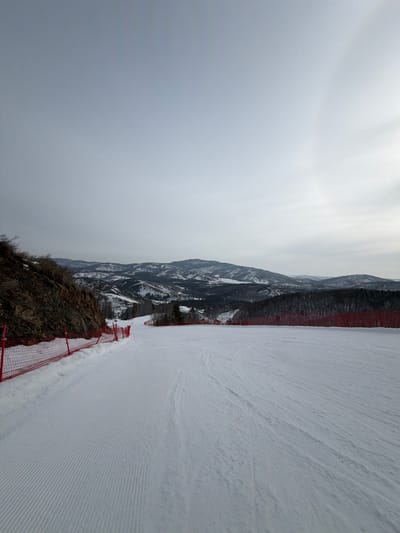 The image displays a snow-covered ski slope located in Novoabzakovo, Russia, under an overcast sky. A faint, partial solar halo is discernible in the upper right quadrant of the sky. The foreground features a wide, groomed snow surface with distinct parallel ridged tracks. Along the left edge of the slope, a red safety netting runs parallel to a steep, rocky embankment containing sparse dry vegetation. A similar red safety netting is visible along the right edge of the slope, beyond which lies a line of snow-dusted coniferous trees. The background reveals multiple layers of snow-covered mountains and hills, some partially forested, receding into the distance under a hazy sky. No individuals, animals, or active equipment are present in the scene.
