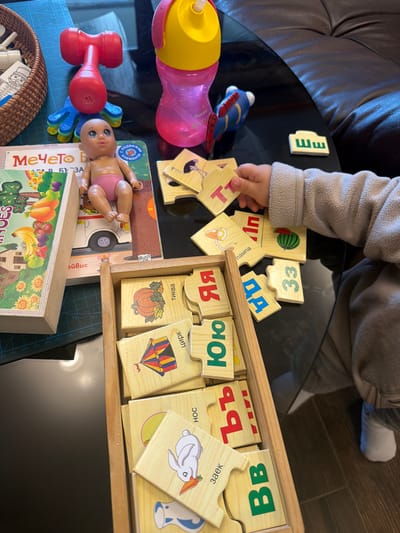 A toddler in grey clothing sits at a black glass table, playing with wooden puzzle pieces and other toys. The puzzle pieces, with images of objects and Cyrillic letters, are scattered near a wooden box containing more puzzle pieces. A baby doll lies next to a book with a fruit illustration on its cover. A red and blue toy hammer, a pink and yellow bottle, and a wicker basket are also on the table. The OCR text indicates the puzzles are related to Bulgarian words. The location is inferred to be related to the Cyrillic script used, potentially within Bulgaria, but the specific city of Кирково is not definitively confirmed from the image itself.
