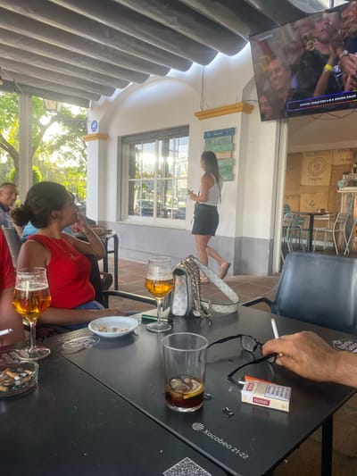 The image depicts an outdoor seating area of a cafe in San Pedro De Alcántara, Spain. A woman with dark hair pulled back is seated at a black table, wearing a red sleeveless top and smoking. In front of her are two glasses of beer, an ashtray filled with cigarette butts, and a small dish of nuts. To the right, a person's hand is reaching for a pair of glasses and a pack of Nobel cigarettes on the table, next to a glass containing a dark beverage with ice and a lemon slice. A lit cigarette is held between the fingers of the hand. In the background, a television screen shows a soccer match with Spanish commentary. Further back, a woman in a white tank top and dark skirt walks past a wall decorated with signs, including one indicating "No tenemos WIFI" (We don't have WIFI). The environment appears to be a sunny day, with trees visible in the distance. The cafe has a covered patio with visible beams.