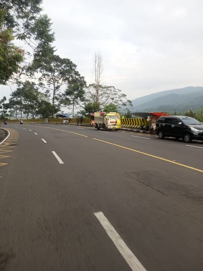 A daytime shot of a curved two-lane road in Taunggyi, Myanmar. The road is asphalt with white dashed lane markers and a solid yellow centerline. A black sedan and a tan pickup truck are parked on the right side of the road, along with a roadside stall. A yellow and black striped barrier runs along the edge of the road. Trees line the left side of the road, and mountains are visible in the background. The sky is overcast. There is a motorcyclist on the left side of the road.