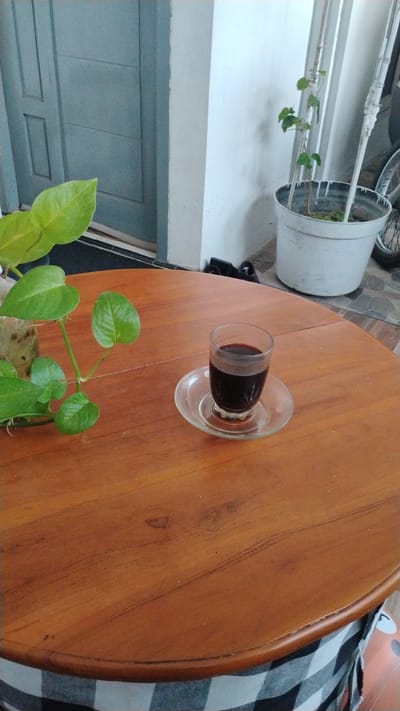 A close-up, high-angle shot shows a small glass of dark liquid, likely coffee, resting on a clear glass saucer on a round, wooden table. To the left of the glass, a potted plant with large, green leaves trails onto the table. In the background, a pale blue door with horizontal paneling is visible. To the right of the door, a white wall with vertical elements is present, along with a large white pot containing a small plant. A bicycle wheel is partially visible behind the white pot. The table is draped with a black and white checkered fabric along its edge. The scene appears to be an outdoor or semi-outdoor setting in Banjarsari, Indonesia.