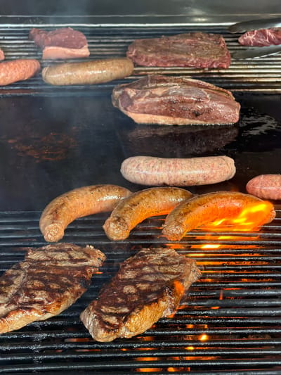 The image shows a grill with various meats and sausages being cooked. There are several pieces of steak, some with visible grill marks, and an assortment of sausages, including what appears to be a lighter-colored type and some that are browning and even flaming. The grill is made of black metal bars. The image was taken in Kerobokan, Indonesia.
