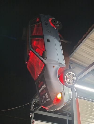 The image depicts a car suspended upside down at a car wash in Șoldănești, Moldova. The car, a light gray hatchback, is positioned with its roof facing downwards and its wheels facing upwards. Red lights emanate from within the car's interior and from the wheel wells, creating a striking visual effect. A sign for "CAR WASH UNTILA" is visible on the side of the car, with text indicating prices or services. The car wash appears to be an outdoor facility, with a corrugated metal roof structure visible in the background. The overall scene is illuminated by artificial lighting, and the sky is dark, suggesting it is nighttime. The car is mounted on a metal support structure.