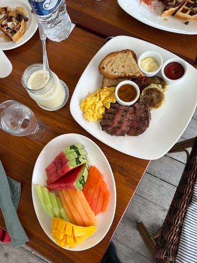 An overhead shot shows a breakfast spread on a wooden table. In the foreground, a white plate holds slices of watermelon, cantaloupe, papaya, and mango. To the left, a glass of water sits next to a green napkin. Above this, a glass jar contains a light-colored smoothie, with a plastic bottle of Crystal Clear water being poured into it. Further up, a plate of waffles with toppings and syrup is partially visible. To the right, another white plate holds scrambled eggs, sliced steak, hash browns, butter, and ketchup. The scene is set outdoors on a wooden deck, with a woven chair visible in the lower right corner. This breakfast is being served at a restaurant in San Pedro Town, Belize.