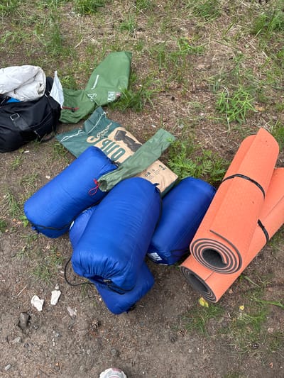 The image depicts camping gear laid out on a patch of ground covered with dry grass and dirt. In the foreground, there are two rolled-up orange sleeping mats with black straps. To the left of the mats, there are at least three rolled-up blue sleeping bags. Behind the sleeping bags, there is a green tent bag. Further back and to the left, there is a black duffel bag with a white folded item on top of it. The environment appears to be outdoors, possibly a campsite. The scene does not contain any people. The location is identified as being in the city of Bolshaya Izhora, Russia.