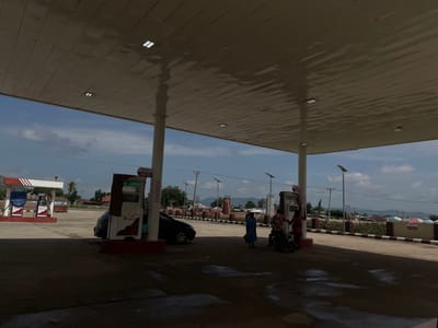 This is an outdoor scene at a Shell gas station in Bassa, Nigeria. The image is oriented sideways. In the foreground, there is a large, flat, light brown roof with several square lights embedded in it. The roof reflects the sky. In the middle ground, there are two Shell-branded gas pumps. One pump has a blue bag on it, and a black car is parked next to it. Two people, one wearing a blue dress and carrying a bag, and another person in an orange and patterned shirt, are standing near the pumps. The other gas pump appears to be unused. In the background, there are trees, buildings with red and white striped accents, and mountains in the distance under a cloudy blue sky. The overall impression is a normal day at a gas station. There is no visible text in the image.