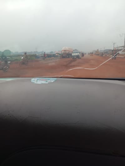 The image depicts a street scene in Imilike, Nigeria, characterized by a dusty, reddish-brown dirt road. The sky is overcast, contributing to a muted, hazy atmosphere. In the foreground, the dashboard of a vehicle is visible, with a reflection or sticker on the windshield.

Further down the road, there is traffic. Several vehicles, including trucks and motorcycles, are present. One white truck with a tank attachment is prominent. People are seen walking and riding motorcycles. The environment appears somewhat undeveloped, with what look like makeshift structures or stalls lining the side of the road. The overall impression is of a busy, dusty road in a possibly rural or semi-urban area of Imilike, Nigeria.