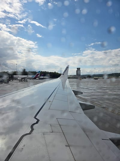This is a ground-level photograph taken from the perspective of an airplane passenger looking out the window. The foreground is dominated by the wing of the aircraft, with visible rivets and panel lines, and water droplets on its surface. To the left, another aircraft is visible, identified by the "Eurowings" logo, parked at a gate with ground crew and equipment around it. Further in the background, other aircraft are seen on the tarmac, one with a Ryanair logo on its tail. The airport tarmac is wet, suggesting recent rain, and the sky is partly cloudy with blue patches. In the distance, a control tower is visible, along with trees and rolling hills, indicating this is an airport in a more rural or suburban setting. The scene captures a moment at an airport before or after a flight, with aircraft and airport infrastructure clearly identifiable. The location is identified as Balice, Poland, which is the location of John Paul II International Airport Kraków–Balice (KRK).