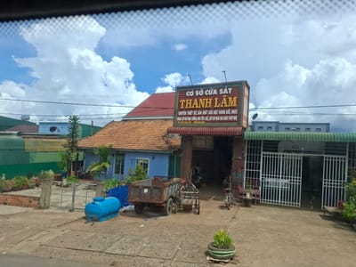 The image shows a street view of a building with a sign that reads "CƠ SỞ CỬA SẮT THÀNH LÂM" in Vietnamese, indicating a metal gate and door workshop. The building is of moderate size with an orange tiled roof and a blue-painted facade. In front of the building, there is a workshop area with a rusty truck and a small trailer. A blue water tank is located on the left side of the scene. A white metal fence with a gate is visible in front of a property next to the workshop. The area appears to be a commercial or industrial zone. The sky is partly cloudy.