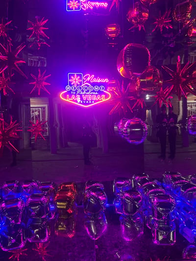 The image shows a reflective surface, possibly a mirrored ceiling, adorned with decorations. There are several star-shaped balloons in red and gold, along with cube-shaped silver and blue balloons. A neon sign that reads "Maison" and "Welcome to Fabulous Las Vegas Nevada" is prominently displayed. The image also features reflections of people standing in the room, though their features are not distinct. The overall lighting creates a purple and blue ambiance. There is no indication that this image is in Ezeiza, Argentina.