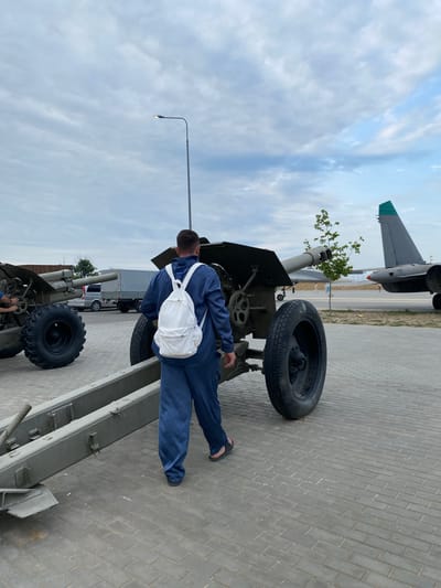 In Arablyar, Russia, a person wearing a dark blue jumpsuit and a white backpack walks away from the camera on a paved area. The person is positioned between two large, green artillery pieces. The artillery pieces are of military design, with large wheels and cannons. In the background, a gray truck with a tarped cargo bed is visible, as are two aircraft, one with a distinctive green tail fin. The sky is overcast with scattered clouds.