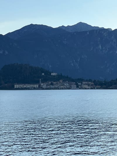 A scenic view showcases a tall, dark mountain range against a light sky. Adjacent to the mountain is a small town with closely packed buildings lining the shore. The right side of the image features water, its surface textured with ripples and reflections, suggesting a slight breeze. The composition offers a blend of natural and urban elements.