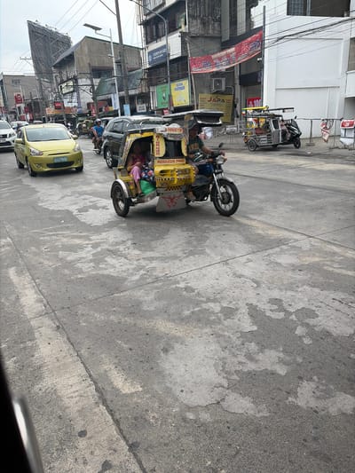 This is a medium-angle, eye-level shot captured on a street in Dagupan City, Philippines. The scene depicts a bustling urban environment under an overcast sky. In the foreground, a weathered concrete road takes up much of the frame, showing signs of wear and tear with patches of lighter concrete and darker asphalt.
Dominating the mid-ground is a yellow and white three-wheeled motorized tricycle. It has a covered passenger compartment, with two individuals, one possibly a child wearing a pink shirt and patterned leggings, and another adult wearing a pink shirt, seated inside. The driver, wearing a blue shirt and white helmet, is operating the vehicle. The tricycle has a license plate that is partially obscured but appears to read "AHN255".
To the left of the tricycle, a bright yellow Mitsubishi Mirage sedan is visible, its front end facing the camera. It has a license plate that reads "AHN255". Further left, another white vehicle is partially visible. Behind the tricycle, a black SUV is also present.
The background is filled with a mix of commercial buildings and structures. A large, angular billboard is prominently displayed on the left. Several signs and banners are visible on the buildings, including one that reads "JAPA" in red and white letters, and another yellow sign with the same logo. A sign for "CHURCH OF GOD BIBLE STUDY CENTER" is also visible. Power lines crisscross the sky, indicating a developed urban setting.
The overall impression is one of a typical street scene in a busy Philippine city, with local transportation and commerce being the central elements.