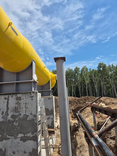 A low-angle, full shot captures a large yellow pipe supported by gray metal beams, elevated on a concrete foundation. The pipe angles upwards towards the left, set against a bright blue sky with scattered white clouds. A tall, gray cylindrical structure stands to the right of the pipe, partially obscuring a dense forest of deciduous trees in the background. In the foreground, to the right of the concrete foundation, is a trench with dirt piles and pipes. A silver ladder leans against the concrete wall. The scene appears to be an outdoor construction site in the Taezhnoe, Russia area, under clear daylight conditions. No people are visible in the frame.