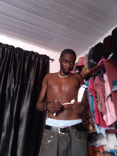 A shirtless Black male stands in what appears to be a cluttered domestic space, holding a toothbrush and a tube of toothpaste. He is wearing dark grey baggy jeans with a black belt and white boxer briefs are visible above the waistband. He has braided hair and is wearing a necklace made of shell beads. To his left, a black curtain is drawn. To his right, a shelf unit filled with clothing and other items is visible. The ceiling is made of white, paneled material. The overall impression is of a casual, personal moment captured in a somewhat disorganized environment, possibly a bedroom or a dressing area. The image does not provide specific information that would allow for a definitive location within Kassa Market, Nigeria.