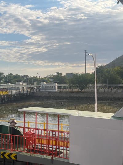 The image captures a waterfront scene in Upt Tambak Sari, Indonesia, under a partly cloudy sky. In the foreground, the edge of a vessel is visible, featuring a green hull, a red railing with yellow accents, and a white structure. A thick rope is coiled around a bollard on the deck.

Beyond the vessel, a body of water separates it from the shore. The shoreline is rocky and appears to be partially exposed due to low tide, with several people scattered across it, some appearing to be foraging or working.

A pier extends into the water, lined with buildings and structures. One building prominently displays the text "SELAMAT DATANG DI TANAH SAMAWA," a welcome message in Indonesian. The area along the shore features a stone wall, a walkway, and several trees and vegetation. In the background, a hillside covered in trees rises, with a mountain visible in the distance. A tall street light stands near the water's edge, and utility poles are also present. The overall atmosphere suggests a functional port or harbor area.
