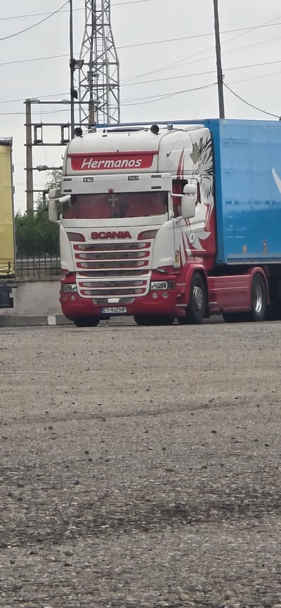 This is an image of a red and white Scania truck with a blue trailer. The truck has "Hermanos" written in red on the side of the cab, and "SCANIA" is visible on the grille. A license plate reading "CT-94 EMP" is also visible. The truck is parked next to a grey, textured surface, possibly a road or wall. In the background, there are power lines and a metal structure, suggesting an industrial or transportation hub setting. The sky is overcast, indicating cloudy weather. The image is taken from a side angle, with the truck's front facing left and the trailer extending to the right. The overall impression is of a commercial vehicle in a somewhat utilitarian environment. There are no people visible in the image. The time of day is indeterminate due to the overcast conditions. The location context of Épila, Spain, does not provide specific visual cues within the image itself.