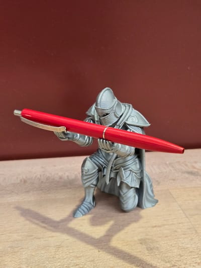 A 3D-printed figurine of a knight in silver armor is kneeling on a wooden surface, holding a red pen horizontally in front of its body with both hands. The knight's helmet is closed, and its left leg is forward with the foot flat on the surface, while its right leg is bent at the knee and tucked behind it. The knight is wearing a cape that drapes down its back. The background is a plain, dark red wall. The scene is lit from the front, casting a shadow of the knight and the pen onto the wooden surface to the left. This image does not contain information about the city of Ansbach, Germany.