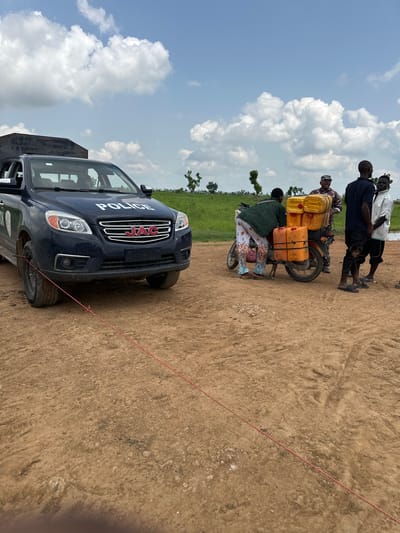 This media file depicts a scene in Garin Dogo, Nigeria. The primary subjects are a group of people and a dark blue police vehicle. One group of men, dressed in casual clothing, stands near a motorcycle laden with several yellow and orange plastic containers. Another person, possibly a woman, is seated on the motorcycle, wearing a green garment and patterned trousers. The police vehicle, a JAC SUV, is parked in the foreground and has the word "POLICE" prominently displayed on its front grill. The scene appears to be outdoors, with a dirt road and a background of green fields and trees under a sky with scattered white clouds. The time of day is likely daytime, given the bright sky and shadows. The overall impression is one of a community engaged in transportation, possibly related to the movement of goods or supplies, with the presence of law enforcement.