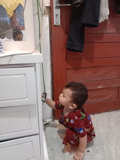 An indoor scene in Lam Ujong, Indonesia, depicts a young child in a dark red, patterned short-sleeved outfit and matching shorts, squatting on a light-colored tiled floor. The child is reaching with their right hand to grasp a black electrical plug connected to a vertical green cable running along a discolored light-colored wall. Adjacent to the child on the left is a white multi-drawer furniture piece. A mirror positioned atop this furniture reflects the upper face of an adult, a portion of a white ceiling fan, and yellow draped fabric. A white container with the partial word "Legion" is also visible on the furniture surface. To the right, a red wooden door with horizontal paneling features a silver handle and a keyhole; dark jeans and a white lace garment hang from its top edge.