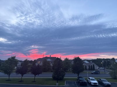 The image captures a sunset scene in Allen Township, United States of America. A vibrant pink and red sky dominates the upper half of the frame, with wispy clouds stretching horizontally across the expanse. Below the sky, a residential area is visible, featuring houses with pitched roofs and manicured lawns. Several trees, some with green foliage and others with dark purple leaves, are planted along a paved road. In the foreground, a parking lot is occupied by four vehicles: a black SUV, a white SUV, a dark grey sedan, and another dark-colored vehicle partially visible. The overall atmosphere suggests a peaceful evening in a suburban setting.
