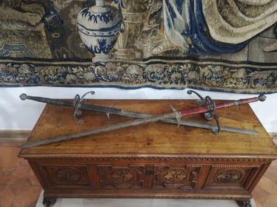 Three antique swords are displayed on a dark wooden chest in an interior room of the Mir Castle Complex in Мірскі сельскі Савет, Belarus. The chest features carved paneling with circular rosette motifs and a beaded border along its top edge. Two straight-bladed swords, possibly rapiers, with ornate metal hilts and wrapped grips (one black, one red), are positioned parallel to each other on the chest. A third sword with a wavy, flamberge-style blade and a simpler hilt is laid diagonally across the other two. The blades show varying degrees of age and discoloration. Behind the chest, a large, multicolored tapestry hangs on a white wall. The tapestry depicts a classical scene, including a blue and white urn, flowing drapery in blue and beige, and a decorative floral border along its lower edge. The floor is partially visible, showing terracotta-colored tiles. No individuals are present.