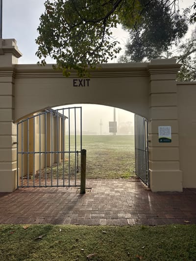 An image captures an archway serving as an exit to a vast, open grassy field, likely a sports oval in Bassendean, Australia. The archway is constructed from light-colored masonry, featuring decorative vertical ridges on its columns. The word "EXIT" is prominently painted vertically in dark blue or purple on the inner left column.

A metal gate, made of horizontal bars, is positioned within the archway. It appears to be open, secured by a chain to a sturdy wooden post on the right side of the passage. The ground directly beneath the arch and extending into the foreground is paved with dark bricks laid in a herringbone pattern. Adjacent to the pathway on the right, a strip of vibrant green, dewy grass is visible, suggesting morning or a damp environment.

Attached to the inner right side of the archway is a rectangular sign with a green border. The white text on a dark background reads: "Please note, in order to ensure the enjoyment of this oval and its facilities, access is permitted from 8am to 8pm daily. On Fridays from 4pm to 8pm during football season, the Bassendean Football Club have priority use of this oval for their junior program and for games on Saturday until the conclusion of the games." A logo, possibly a green shield with a tree, is visible below the text.

Beyond the archway, the expansive grassy field extends into a soft mist or fog, obscuring the distant horizon and creating a serene, somewhat mysterious atmosphere. Faint structures, possibly goalposts or light towers, are barely discernible through the haze. Overhanging the left side of the archway are branches of a leafy tree, its foliage silhouetted against the bright, overcast sky. The scene depicts a quiet moment of transition, with no people visible, and the damp appearance of the grass and fog suggesting either early morning or a period of moisture.