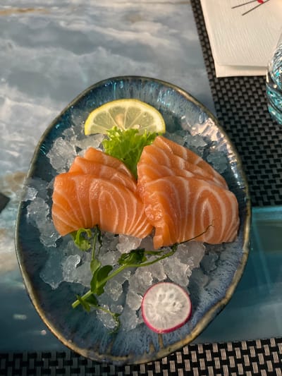 An overhead shot captures a beautifully arranged dish of salmon sashimi served on a bed of crushed ice. The main subject is several thick slices of vibrant orange salmon, artfully fanned out in two separate groupings on a rustic-style blueish ceramic plate with an irregular, dark rim.

Accompanying the salmon are garnishes including a bright yellow lemon wedge, a small cluster of fresh green lettuce, and a thin slice of red and white radish. A few delicate green pea shoots or similar tendrils are also scattered on the ice, adding a touch of freshness. The crushed ice keeps the fish chilled and enhances its visual appeal.

The plate rests on a reflective, light-colored surface, possibly a marble-patterned table, which shows subtle reflections of the dish. To the upper left, a corner of a white napkin is visible, featuring a red graphic that appears to be a stylized representation of chopsticks. A dark grey or black woven placemat is partially visible under the napkin and along the right side of the plate, providing a textured contrast to the smooth table and food. The scene suggests a dining setting, likely in a restaurant specializing in Japanese cuisine in Basiglio, Italy. The lighting is soft and even, highlighting the fresh colors of the food.