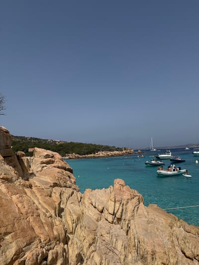 This image depicts a scenic coastal view with a clear blue sky and turquoise water. In the foreground, large, weathered rock formations dominate the landscape, casting shadows and highlighting their rough textures. Beyond the rocks, the crystal-clear water reveals the seabed beneath.
Several boats are anchored in the water, including a white inflatable dinghy with people on board. The individuals in the dinghy appear to be enjoying a day out on the water, with some sitting and others standing. In the background, a lush green coastline stretches along the water's edge, suggesting a Mediterranean setting.
The lighting indicates it is daytime, likely a sunny day given the bright illumination and clear skies. The presence of boats and people enjoying the water suggests a leisurely activity, possibly swimming or exploring the coastline. The overall mood of the image is serene and inviting, capturing the beauty of a summer day by the sea. The location is likely Palau, Italy, as suggested by the context. There is no visible text in the image.