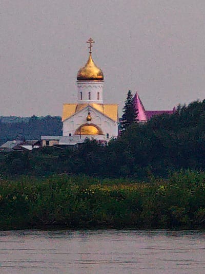 An Orthodox church with a golden dome and a gold cross on top stands prominently against a hazy, overcast sky. The white church has yellow trim and a smaller golden dome. To the right of the main church, a smaller building with a pink-hued roof is visible, partially obscured by dark green trees. In the foreground, a body of water with a reflective surface stretches across the bottom of the frame, bordered by lush green vegetation. The scene appears to be a landscape view of a settlement, possibly the village of Kazachinskoe, Russia, given the architectural style of the church. There are no people or discernible actions captured in the image.