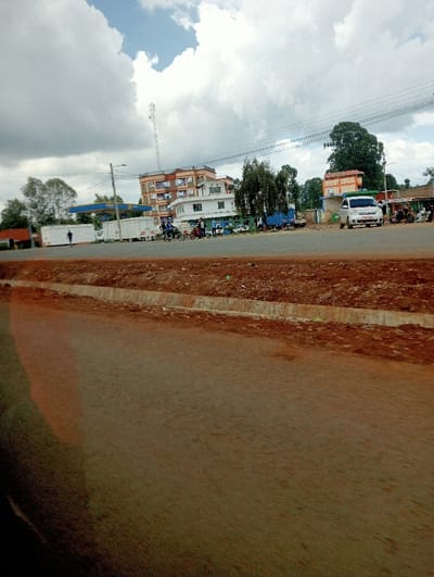A street scene with several buildings and vehicles visible under a cloudy sky. The foreground shows a section of a road. There's a gas station and a tall communication tower. People are gathered near the roadside, and vehicles are parked along the street. Electrical wires stretch across the sky. The buildings vary in height, and some have multiple stories. The overall impression is of a busy, small-town environment.