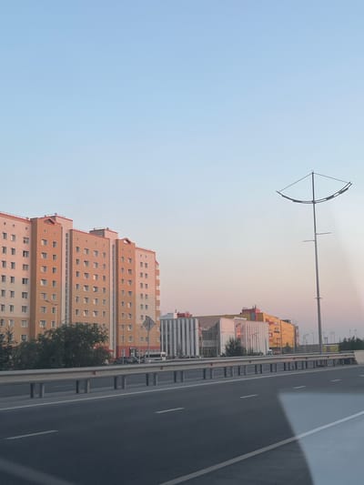The image captures a section of a highway in Novy Urengoy, Russia, with a clear sky and several buildings in the background. On the left, a tall, multi-story apartment building with a predominantly orange and beige facade is visible. It features numerous windows arranged in a grid pattern. To the right of the apartment building, a series of modern commercial buildings with varied architectural styles and colors, including yellow, white, and pink, are present. One of these buildings appears to be a shopping center. A tall, distinctive street lamp with a curved top is positioned in the middle ground to the right. The highway itself is in the foreground, characterized by asphalt lanes marked with white lines and a metal guardrail separating it from the area beyond. A few trees and street signs are also discernible. No people or vehicles are clearly identifiable in action or interaction within the frame. The overall scene suggests a daytime view of an urban or suburban environment under a light blue sky.
