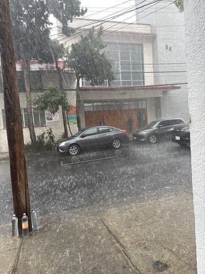 The image depicts a street scene in Tizapan, Mexico, during a heavy rainstorm. The foreground shows a sidewalk with three plastic water bottles standing upright next to a wooden utility pole. The street is wet and reflects the overhead light, with numerous raindrops visible in the air and on the pavement. Several cars are parked on the street, including a dark-colored sedan and an SUV. In the background, a building with a light-colored textured facade and a red-tiled roof is visible. Trees with dense foliage are scattered along the street. Two individuals are partially visible, one standing near a car and another near a doorway. The weather conditions are characterized by intense rainfall, with water obscuring visibility and creating a somber atmosphere.