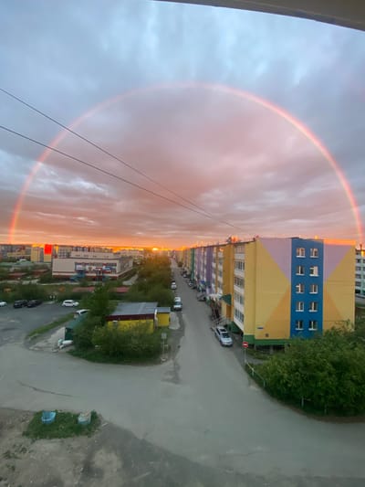 This image depicts an urban scene in Udachny, Russia, featuring a vibrant rainbow arcing across a cloudy sky at sunset. The setting is residential, with several multi-story apartment buildings dominating the landscape. The buildings display a variety of colorful facades, including yellow, blue, pink, and purple sections, with visible windows and balconies. A paved road runs alongside the buildings, with parked cars and some greenery interspersed.
The sun is low in the sky, casting a warm, orange glow on the clouds and the tops of the buildings, indicating it is either sunrise or sunset. The weather appears to be a mix of clouds and clearing skies, with the rainbow suggesting recent or ongoing precipitation. There are power lines running diagonally across the frame, adding to the urban feel. The overall mood is somewhat serene, with the natural phenomenon of the rainbow providing a striking contrast to the man-made structures. No people are clearly visible in the image. The location context of Udachny, Russia, suggests a northern climate, which could make such atmospheric phenomena particularly striking.