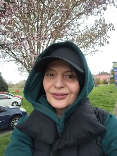 The image depicts a female individual in a selfie-style portrait, situated outdoors in Souffelweyersheim, France. The subject, appearing middle-aged, is looking directly at the camera with a slight smile. She is wearing a dark teal hooded sweatshirt, a black baseball cap positioned under the hood, and a black puffer vest.
The background on the left side of the frame shows a paved area adjacent to a grassy verge. Several vehicles are parked on the paved area, including a blue sedan in the mid-ground, a white car, and a partially visible red vehicle further back. Behind the vehicles and grass, a large tree with light pink blossoms is prominently visible, suggesting a spring season under an overcast sky.
The right side of the background features a wide expanse of green grass dotted with small white flowers. A commercial sign with white and blue elements is partially visible above the grass. In the distance, a two-story building with a light brown or pinkish facade and a darker roof is present, alongside another tree with bare branches displaying catkins or seed pods. The sky throughout the entire scene is uniformly overcast.