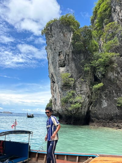 A young man wearing a blue and white striped shirt, sunglasses, and dark pants stands on a wooden boat. He is looking to his left, with a prominent rock cliff face behind him, covered in lush green vegetation. The water is a clear turquoise color. In the background, there are other boats, including a long-tail boat and a larger vessel, and a cloudy blue sky. The scene appears to be a scenic location, possibly near Ton Sai, Thailand, known for its dramatic limestone karsts and clear waters.