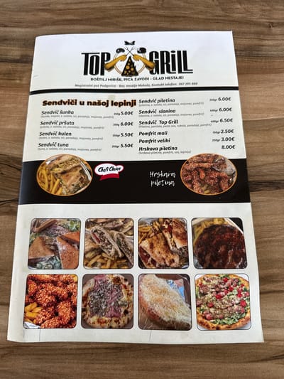 This media file is a photograph of a menu from "Top Grill," a restaurant located in Mojanovići, Montenegro. The menu is presented on a glossy paper, likely as part of a promotional flyer or placemat, placed on a wooden surface.

The menu is divided into sections, showcasing various food items offered by the restaurant. Prominently featured are images of different sandwiches, pizzas, and french fries, presented in a grid layout. The items are described with their ingredients and prices, often in euros. Some specific menu items include "Sendviči u našoj lepinji" (sandwiches in our flatbread), "Sendvič piletina" (chicken sandwich), "Sendvič šunka" (ham sandwich), "Sendvič slanina" (bacon sandwich), "Sendvič pršuta" (prosciutto sandwich), "Sendvič Top Grill," "Sendvič kulen" (Kulen sausage sandwich), "Sendvič tuna" (tuna sandwich), "Pomfrit mali" (small fries), "Pomfrit veliki" (large fries), and "Hrskava piletina" (crispy chicken). The prices range from 2.50€ to 8.00€.

Additional text on the menu provides the restaurant's name, location ("Magistralni put Podgorica - Bar, naselje Mahala"), and contact information, including a phone number. A small "Chef's Choice" badge is also visible. The overall impression is a vibrant and enticing presentation of fast food offerings, designed to attract customers. The lighting suggests an indoor setting, possibly a restaurant or an area where the menu is being displayed. There are no people visible in the image, and the focus is solely on the food and menu information.