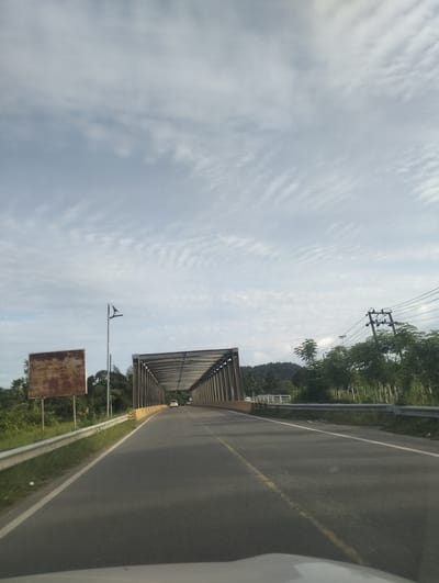 The image shows a road leading towards a metal bridge with a covered walkway. The road is paved with asphalt and has a white line marking the edge and a yellow line marking the center. There are guardrails on both sides of the road. On the left side of the road, there is a rusty, rectangular sign. Further down the road, there are two vehicles. To the right of the road, there are trees and power lines. In the background, there are hills covered in trees. The sky is cloudy. The location is indicated as Babah Dua, Indonesia.